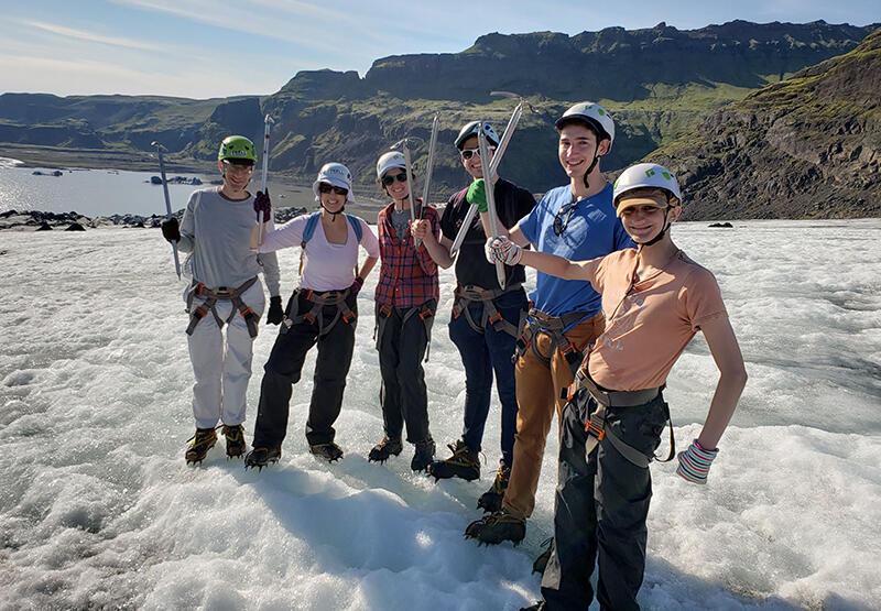Six smiling people in rock climbing gear standing in the snow with hills visible in the background