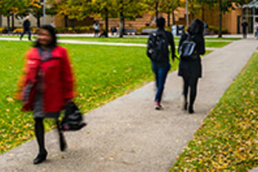 several people walking on a cement walkway on MIT’s Hockfield Court, a grassy plaza surrounded by campus buildings