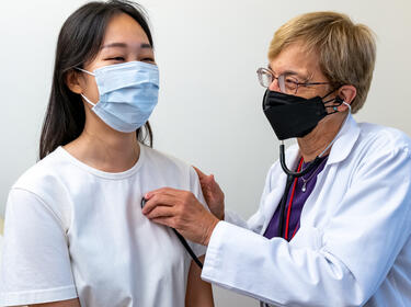 Waist up view of a patient and a clinician, both wearing PPE masks, the clinician, wearing a white coat, listens to the patient’s heart with a stethoscope.