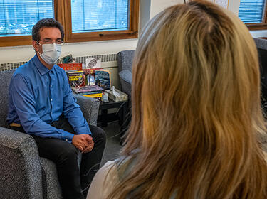 Head and shoulders rear view of a patients head with long brown and blonde streaked hair. A clinician wearing a PPE mask sits in a comfortable armchair while looking toward the patient