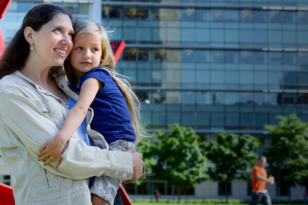 smiling young parent standing outdoors next to a large red sculpture while holding a small child in her arms