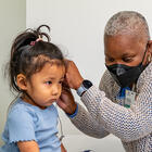 Clinician wearing a PPE mask hold an instrument at the left ear of a toddler-age patient while the patient sits looking forward toward the right side of the frame.